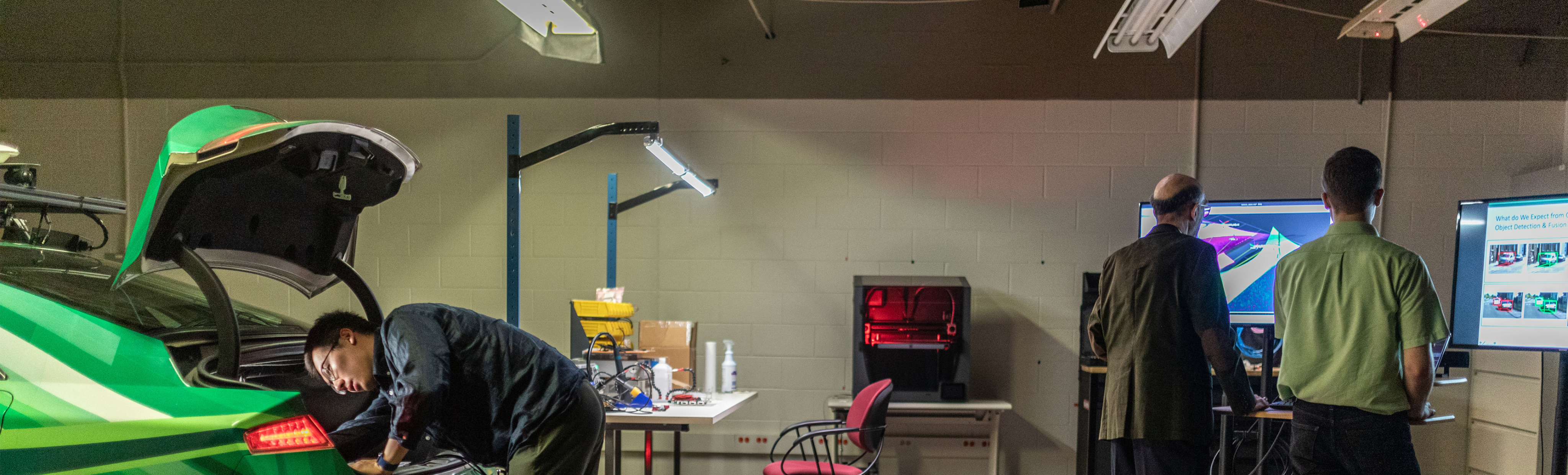 In a research lab, a researcher wearing a dark shirt leans over the back of a bright green car, appearing to work on it. In the background, two other men stand and look at large computer monitors, and a 3D printer is visible on a nearby table.
